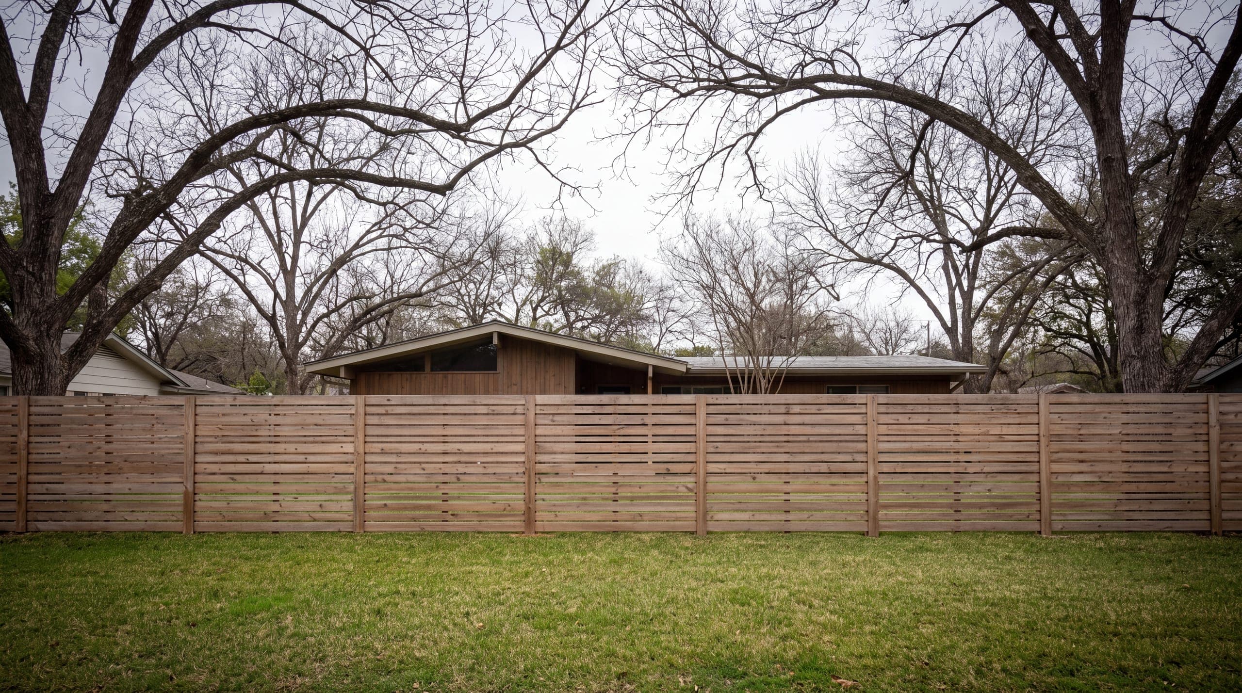 A horizontal cedar slat fence across the front of a low mid-century house under pecans