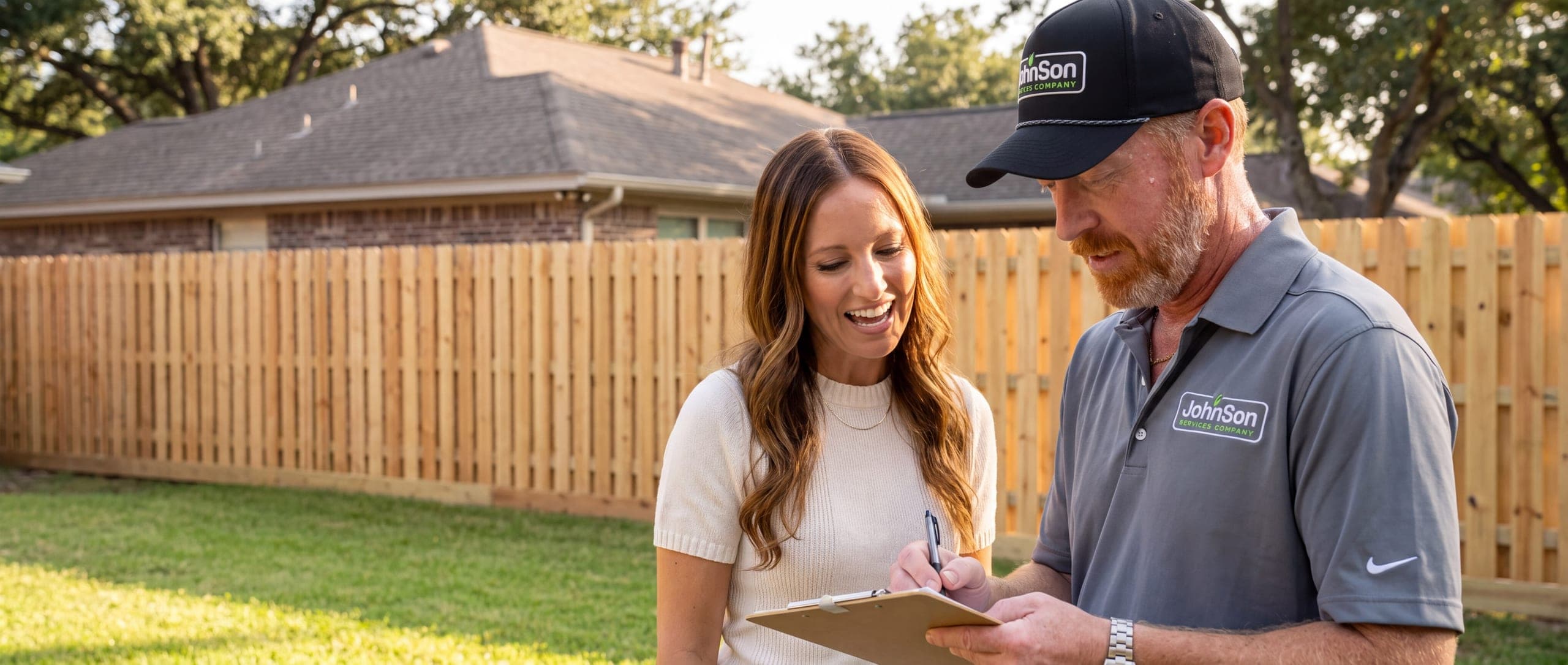 A Johnson estimator writing on a clipboard beside a homeowner in front of a newly built wood fence at an established Texas home