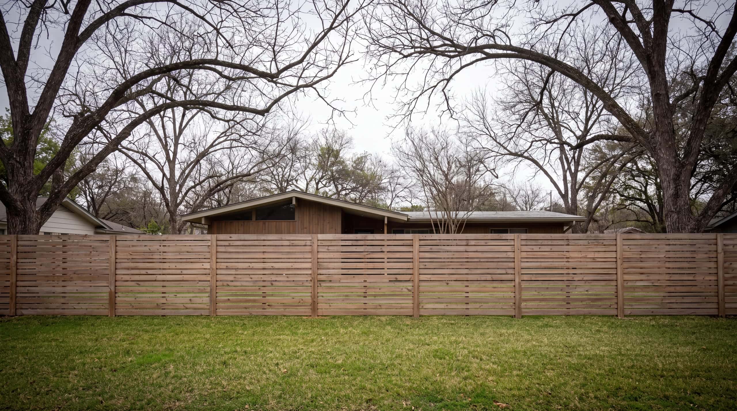A horizontal cedar slat fence across the front of a low mid-century house under pecans